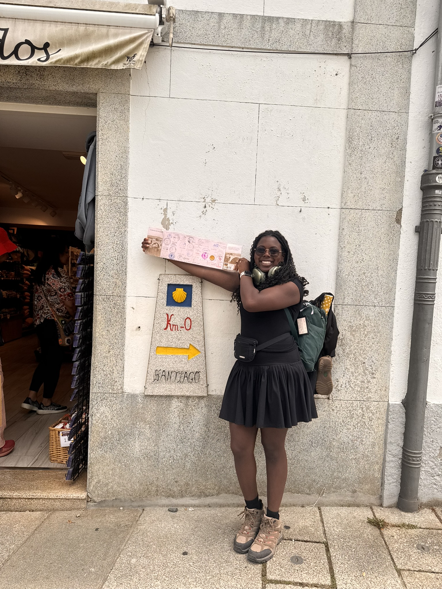 Mesha holds up her Pilgrim Passport in front of a Camino trail marker, a yellow shell on a blue square.