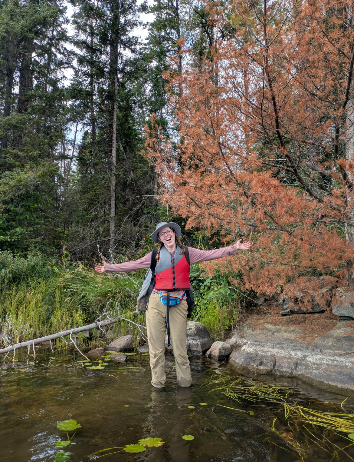 Ava wades in water just off the bank with rocks, trees, and grass around her wearing a sunhat and lifejacket.