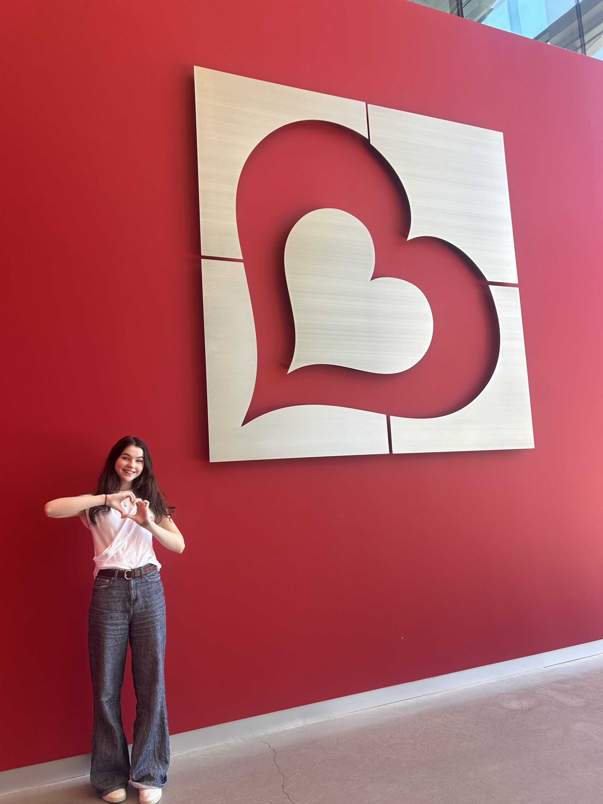 Maylan poses making a heart with her hands in front of a red wall with a large metal Burlington logo on it.